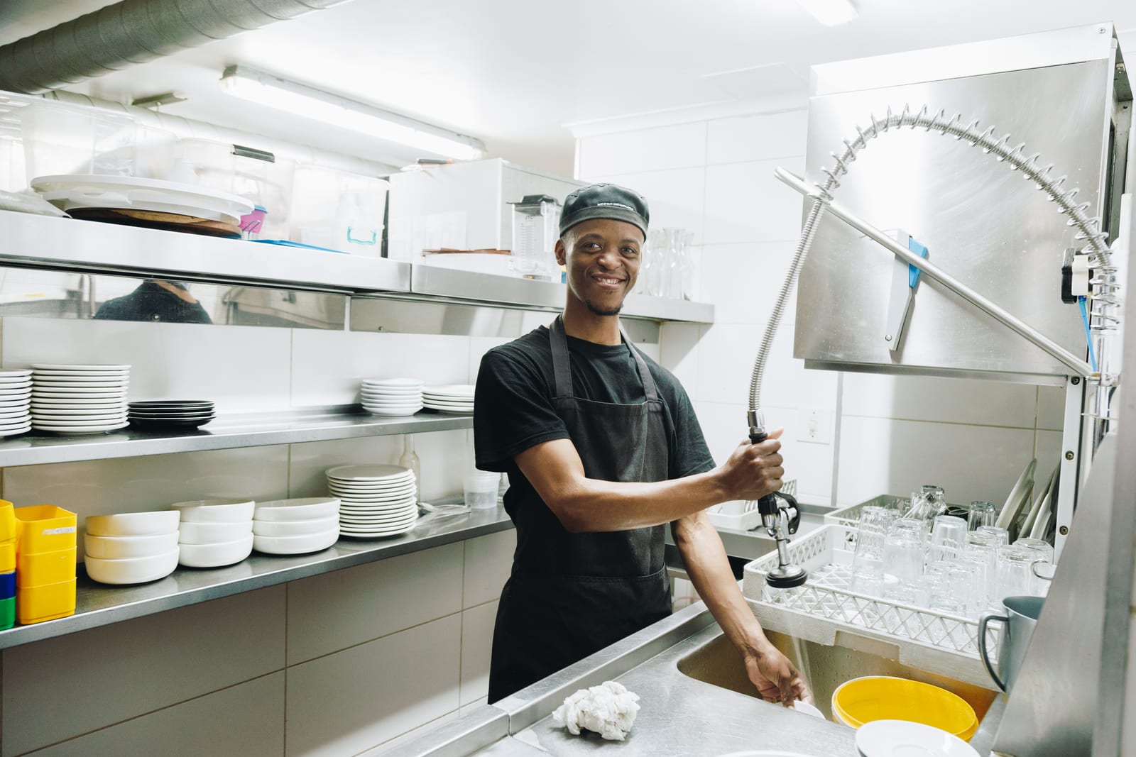 A kitchen worker in a cap and apron smiling at the glass-washing station in a commercial kitchen